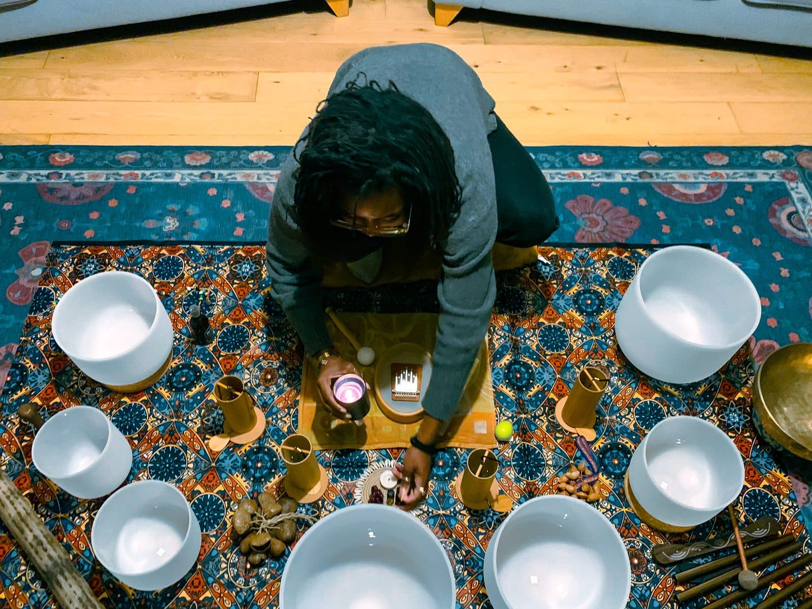 Overhead view of a participant kneeling among singing bowls arranged on a patterned rug during a sound bath